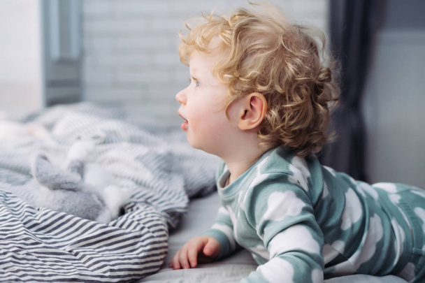 funny little curly baby boy in blue pajamas lies in bed, woke up - Фото, изображение