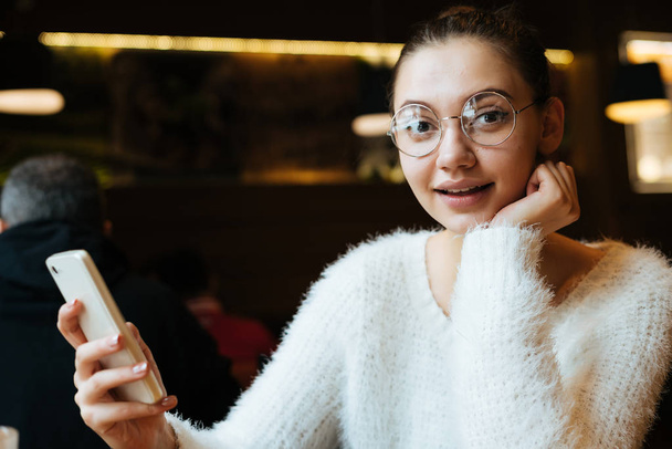 funny young girl student wearing glasses looks surprised, holds smartphone, in cafe - Photo, image