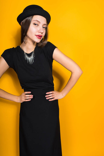 attractive young fashion girl in trendy black dress and hat posing on yellow background - Fotoğraf, Görsel