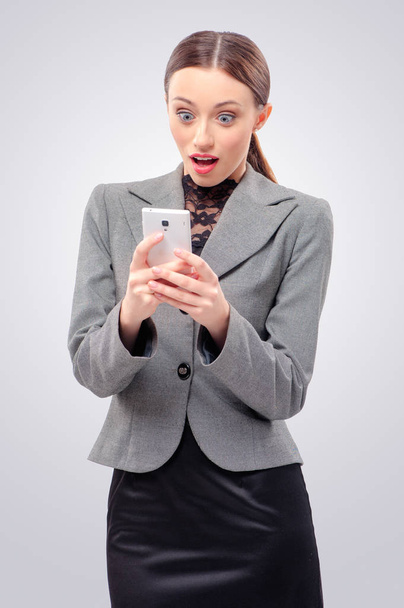 Surprised young caucasian business woman holding mobile phone, looking at it and keeping mouth open while standing against grey background - Foto, Imagen