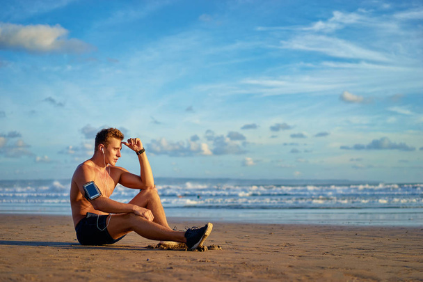 Mladý pohledný muž s sluchátka a smartphone sedí na pláži. - Fotografie, Obrázek