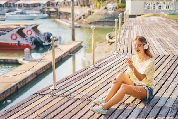 Happy young woman with earphones is listening music while sitting on deck broadwalk in marine harbor. - Valokuva, kuva