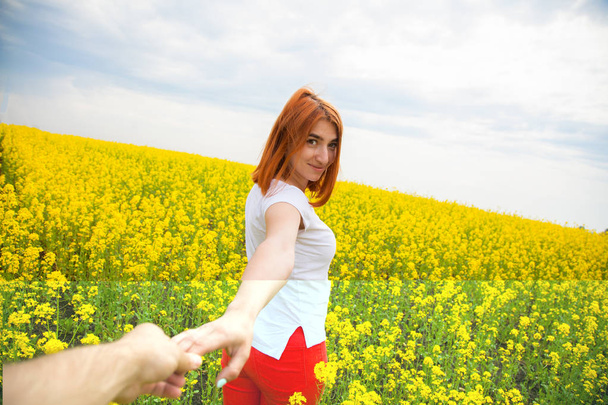 Uma jovem ruiva com uma camiseta branca e jeans vermelhos sorri e leva a mão do homem molhado para as aventuras de um enorme campo de flores amarelas em um dia quente de verão, siga-me
 - Foto, Imagem