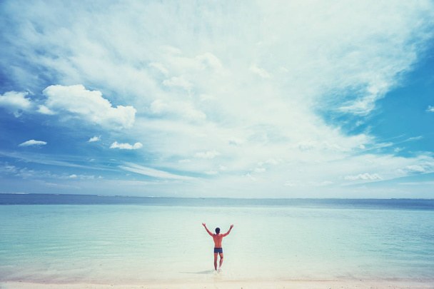 Happy young man rising hands up standing on tropical beach enjoying beautiful view. - Foto, afbeelding