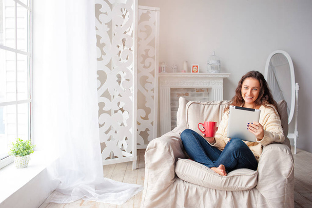 Beautiful young woman with cup of tea using tablet computer while sitting on armchair at home. - Fotografie, Obrázek