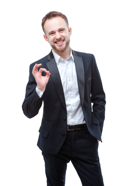 Handsome young bearded man in full suit showing OK sign and smiling while standing isolated on white background - Photo, image