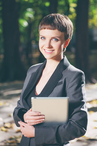 Mulher caucasiana jovem confiante em formalwear segurando tablet digital e sorrindo enquanto está de pé no parque ao ar livre
 - Foto, Imagem