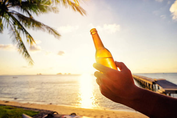  Male hand holding bottle of beer on the sea beach. - Φωτογραφία, εικόνα