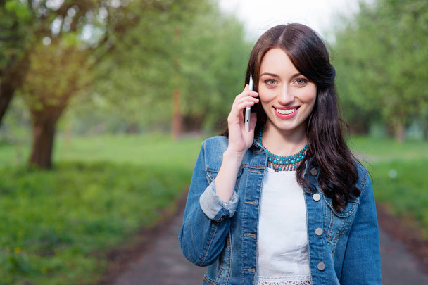 Attractive young woman talking by smartphone while walking by summer park. - 写真・画像