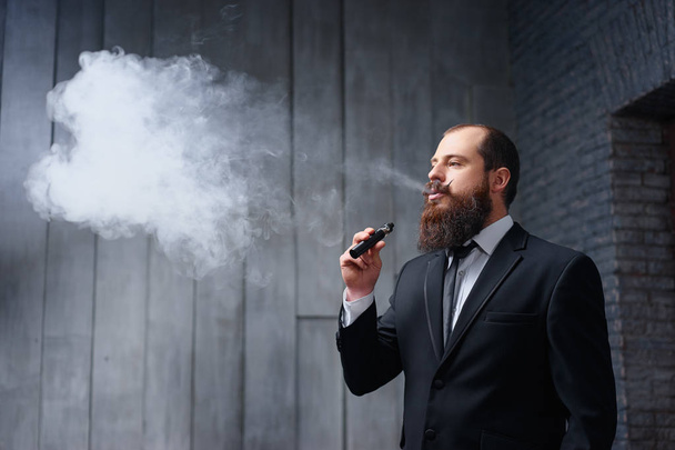 Handsome young bearded man in suit smoking vaporizer electronic cigarette. - Fotó, kép
