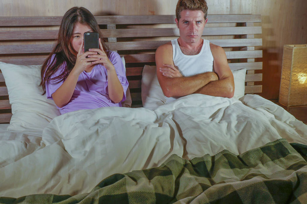 young attractive mixed ethnicity Asian and Caucasian couple in bed with social media addict girlfriend using internet mobile phone ignoring frustrated and upset man moody in relationship problem - Photo, Image