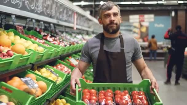 Dolly shot of young salesman in apron carrying box of tomatoes in supermarket in fruit and vegetables department. Salesperson, food store and prosefession concept. - Felvétel, videó