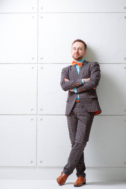Portrait of  young bearded man in plaid suit and bowtie keeping arms crossed and looking at camera while standing against white wall. - Foto, Bild