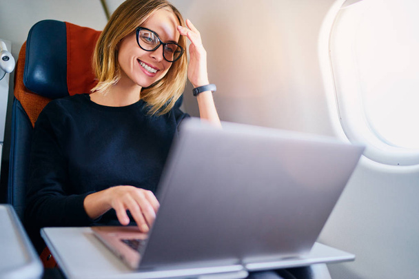 Pretty young business woman working on laptop computer while sitting in airplane. - Photo, image