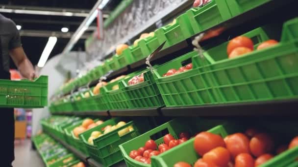 Strong salesman wearing uniform is bringing plastic box of tomatoes and putting on shelf in supermarket in fruit and vegetables department. Containers with food are visible. - Materiał filmowy, wideo