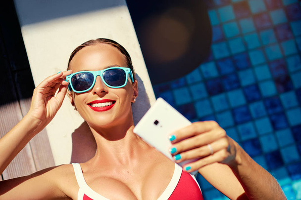 Colorful portrait of pretty young woman using smartphone taking selfie in swimming pool. - Photo, Image