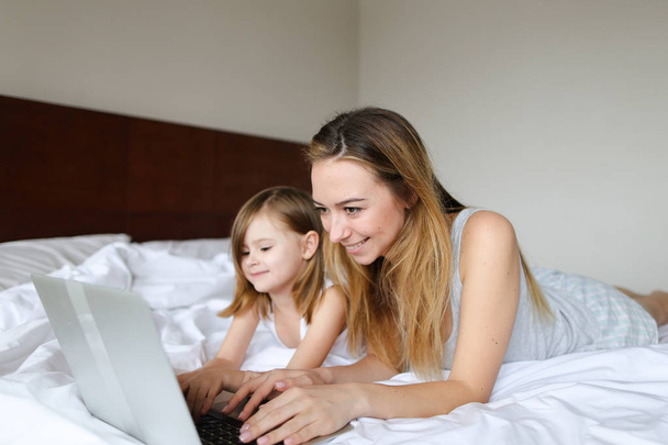 Young smiling woman lying in bed with little daughter in morning and using laptop. - Photo, Image