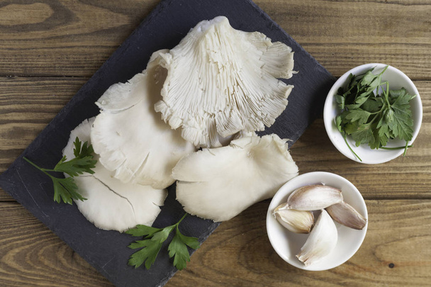 Cogumelos de cardo cru na placa de ardósia, tigela de alho e tigela de salsa na mesa de madeira
 - Foto, Imagem
