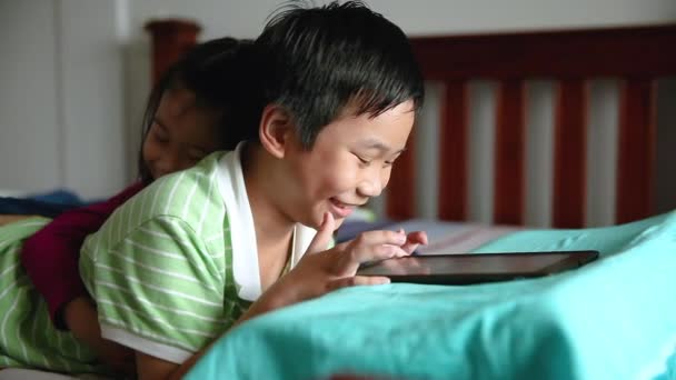 Asian children using digital tablet. Happily sister smiling and cheering her brother near by. Cute boy playing games excitedly on touchpad and lying prone on bed.  - Filmati, video