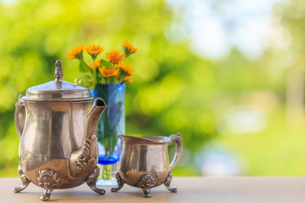 An old beautiful teapot and teacup and yellow flowers in a blue glass pot on wooden board with nature background. - Photo, Image