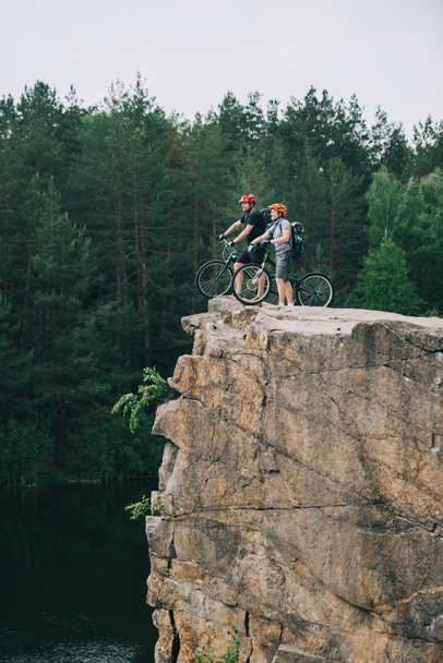 side view of young trial bikers standing on rocky cliff with blurred pine forest on background - Photo, Image