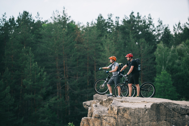 vista lateral de los jóvenes ciclistas de trial de pie sobre un acantilado rocoso con un bosque de pinos borroso en el fondo y apuntando a algún lugar
 - Foto, imagen