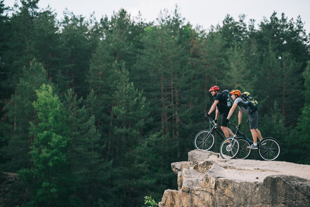 Zijaanzicht van jonge trial bikers op rotsachtige klip met wazig dennenbos op de achtergrond en kijkend naar kant - Foto, afbeelding