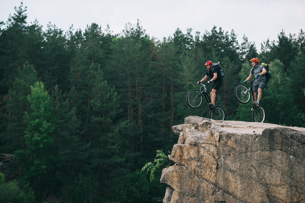 vista lateral de los jóvenes ciclistas de trial con mochilas de pie sobre ruedas traseras en acantilado rocoso con bosque de pino borroso en el fondo
 - Foto, Imagen