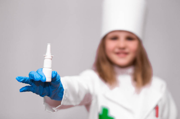 Usmívající se holčička v lékařské uniformě drží nosní sprej nebo kapky pro zdraví v bílé láhvi. Lékařská dívka doporučuje medicínu nos sprej - Fotografie, Obrázek