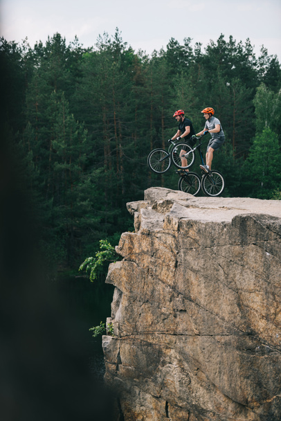 side view of trial bikers standing on back wheels on rocky cliff with blurred pine forest on background - Photo, Image