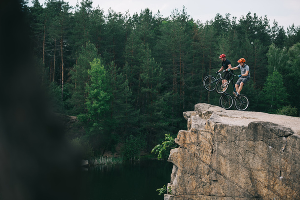 side view of extreme trial bikers standing on back wheels on rocky cliff with blurred pine forest on background - Photo, Image