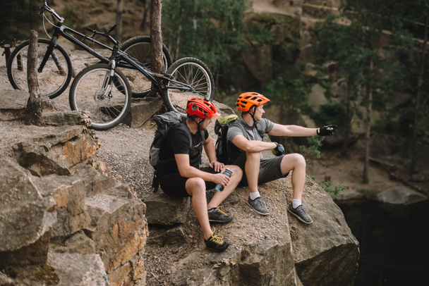 vista de ángulo alto de los jóvenes ciclistas de trial que se relajan en el acantilado rocoso después de montar y mirar hacia otro lado
 - Foto, imagen