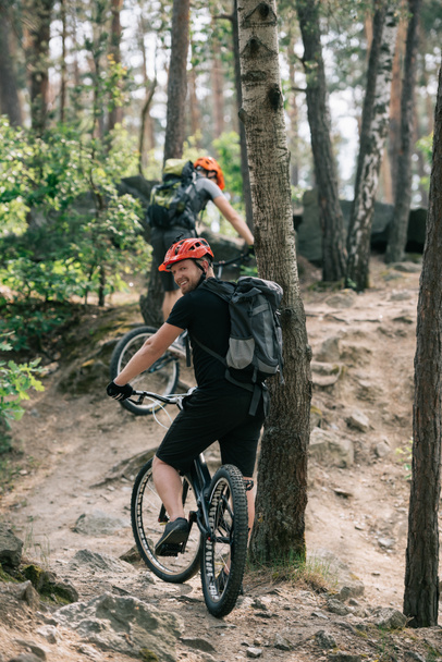 uśmiechnięty mężczyzna ekstremalnych rowerzystów jazda na rowerze górskim z przyjacielem w lesie  - Zdjęcie, obraz