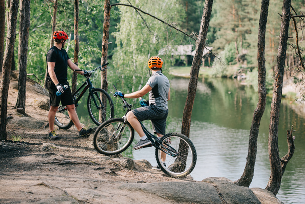 rear view of male extreme cyclists in helmets with mountain bikes resting with sport bottles of water and looking at river in forest  - Photo, Image