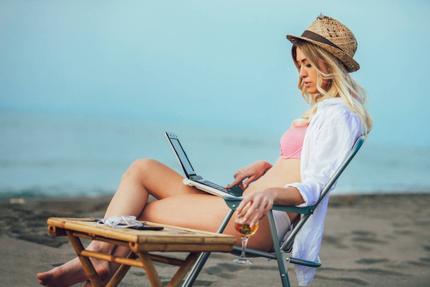 Mujer sexy joven con sombrero usando el ordenador portátil en la playa. Chica Freelancer trabajando
 - Foto, imagen