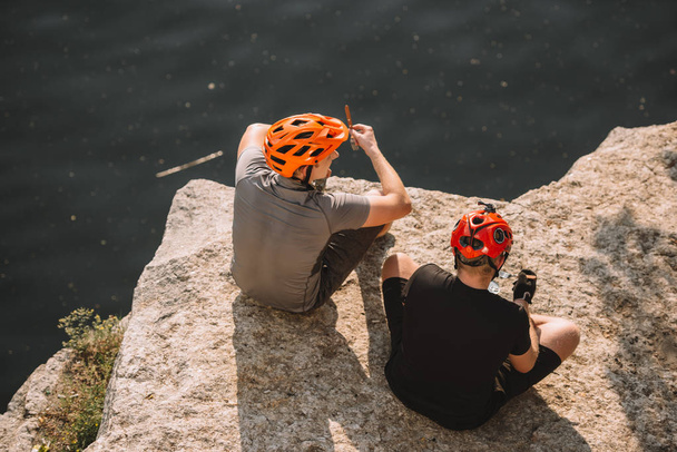 male hikers in protective helmets resting and eating canned food on rocky cliff over river - Fotografie, Obrázek