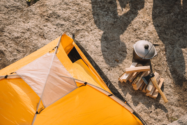 высокий угол обзора туристической палатки, топора, бревна, котла и тени людей на земле
 - Фото, изображение