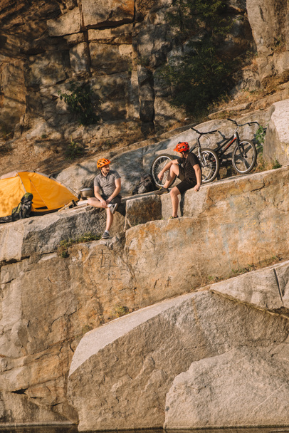 two male travelers in protective helmets resting near tent and bicycles on rocky cliff  - Photo, Image