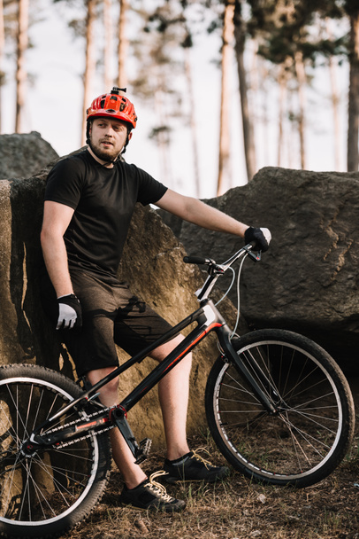 komea nuori kokeiluversio biker nojaa takaisin kiviä ulkona metsässä ja katselee kameraa
 - Valokuva, kuva