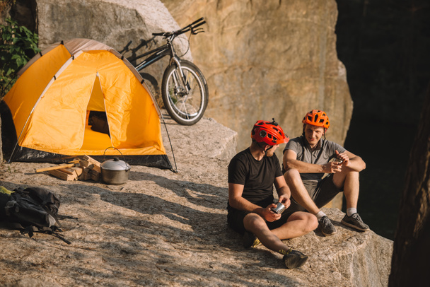 felices jóvenes viajeros en bicicleta con comida enlatada sentados en el acantilado
 - Foto, Imagen