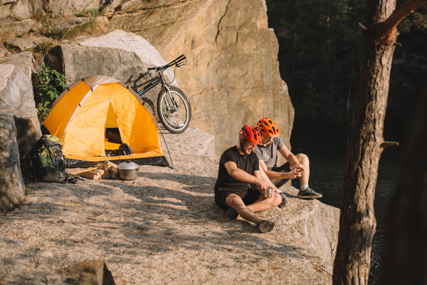 jóvenes ciclistas activos con comida enlatada sentados en el acantilado
 - Foto, Imagen