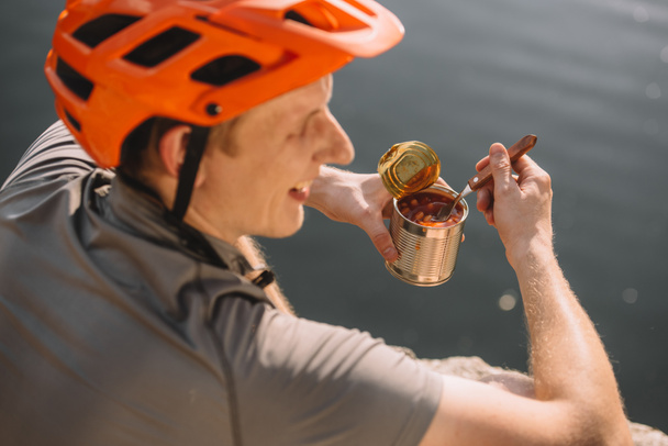 joven viajero guapo en casco comiendo comida enlatada frente al agua
 - Foto, Imagen