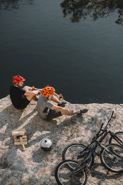 vista de ángulo alto de los jóvenes ciclistas de trial comiendo comida enlatada en un acantilado rocoso
 - Foto, Imagen