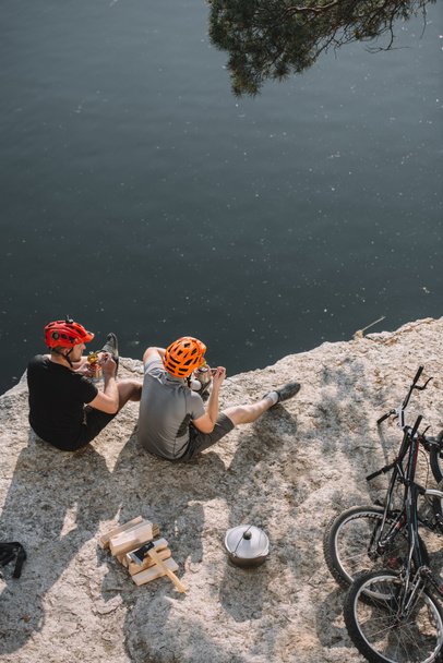 vista de ángulo alto de los viajeros en bicicleta comiendo comida enlatada en un acantilado rocoso sobre el lago
 - Foto, imagen