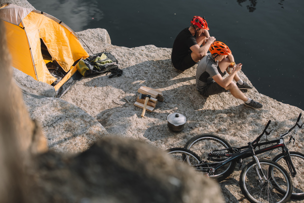vista de ángulo alto de los viajeros activos en bicicleta comiendo comida enlatada en acampada en acantilado rocoso
 - Foto, Imagen