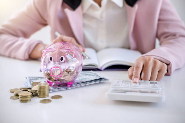 Business woman putting coins into piggy bank, writing to report and using calculator to analysis business investment strategy with income of money coin and dollar, financial concept. - Foto, Imagen
