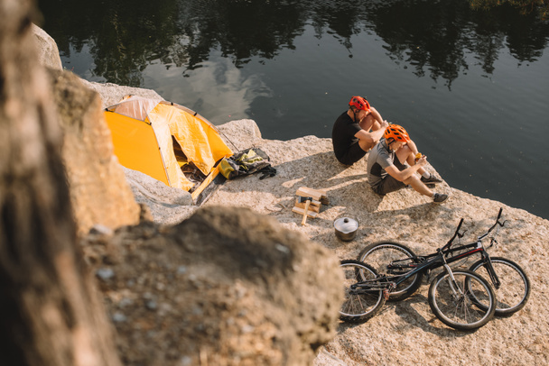 vista de ángulo alto de ciclistas de prueba activos comiendo comida enlatada en acampada en acantilado rocoso
 - Foto, imagen