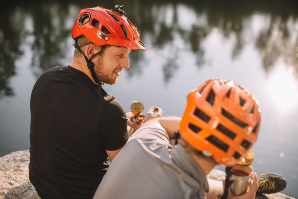 jóvenes viajeros activos que comen alimentos enlatados en el camping frente a la superficie del agua
 - Foto, Imagen
