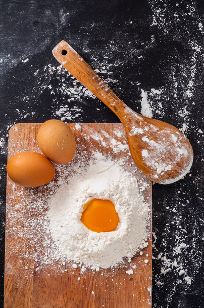 ξύλινο κουτάλι κοντά ένα ωμό αυγό στο σωρό του αλευριού και δύο ολόκληρα αυγά  - Φωτογραφία, εικόνα