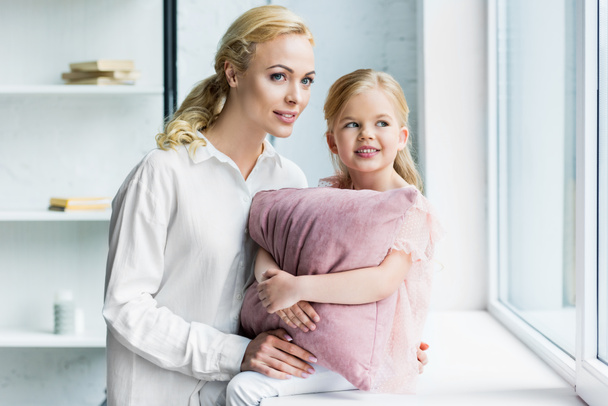 mother and adorable smiling daughter with pillow looking at window at home - Foto, immagini
